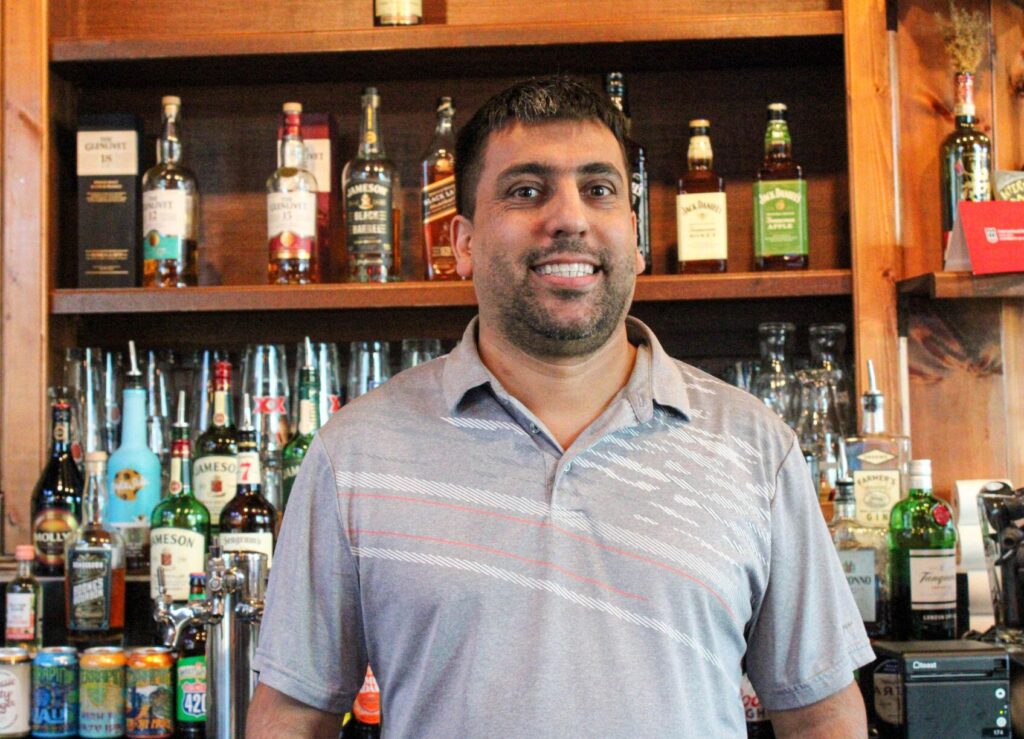 Picture of a man smiling and standing in front of aa bar